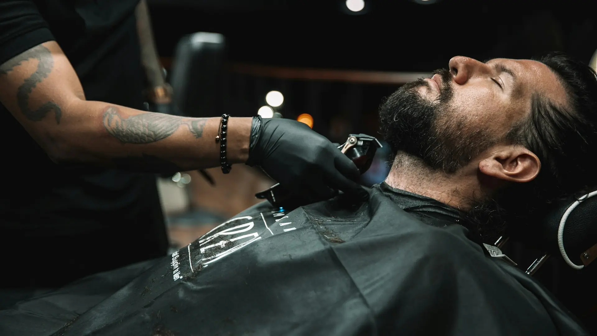 Tattooed barber in black gloves trimming a client's beard with clippers